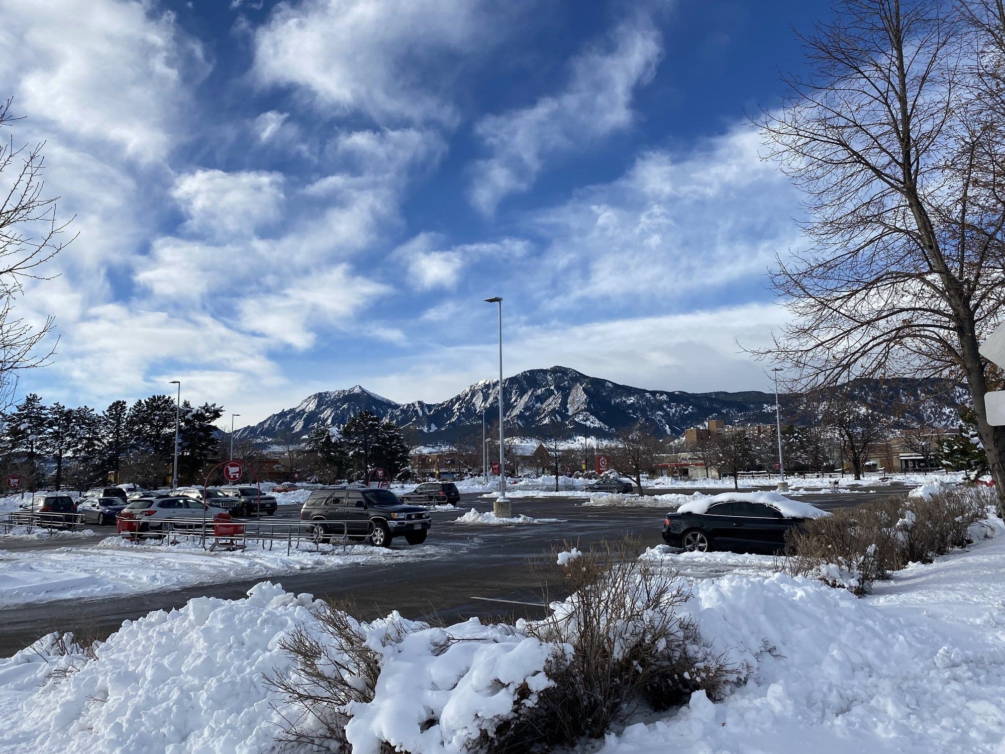 Snowbanks and bare trees in front of the Flatirons under a blue sky full of white clouds