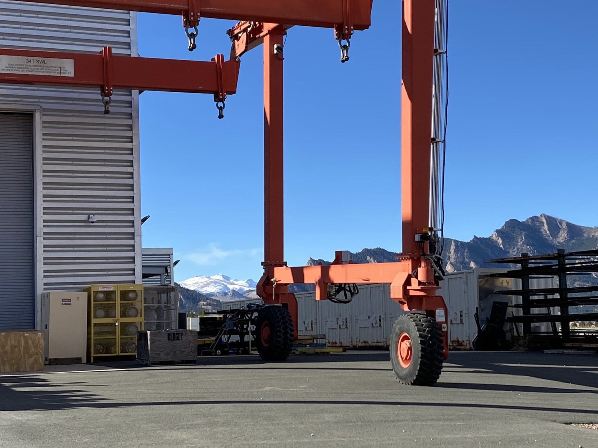 An orange gantry crane in a laboratory yard beside a steel shed, with the Flatirons and a snow-covered peak under a clear blue sky