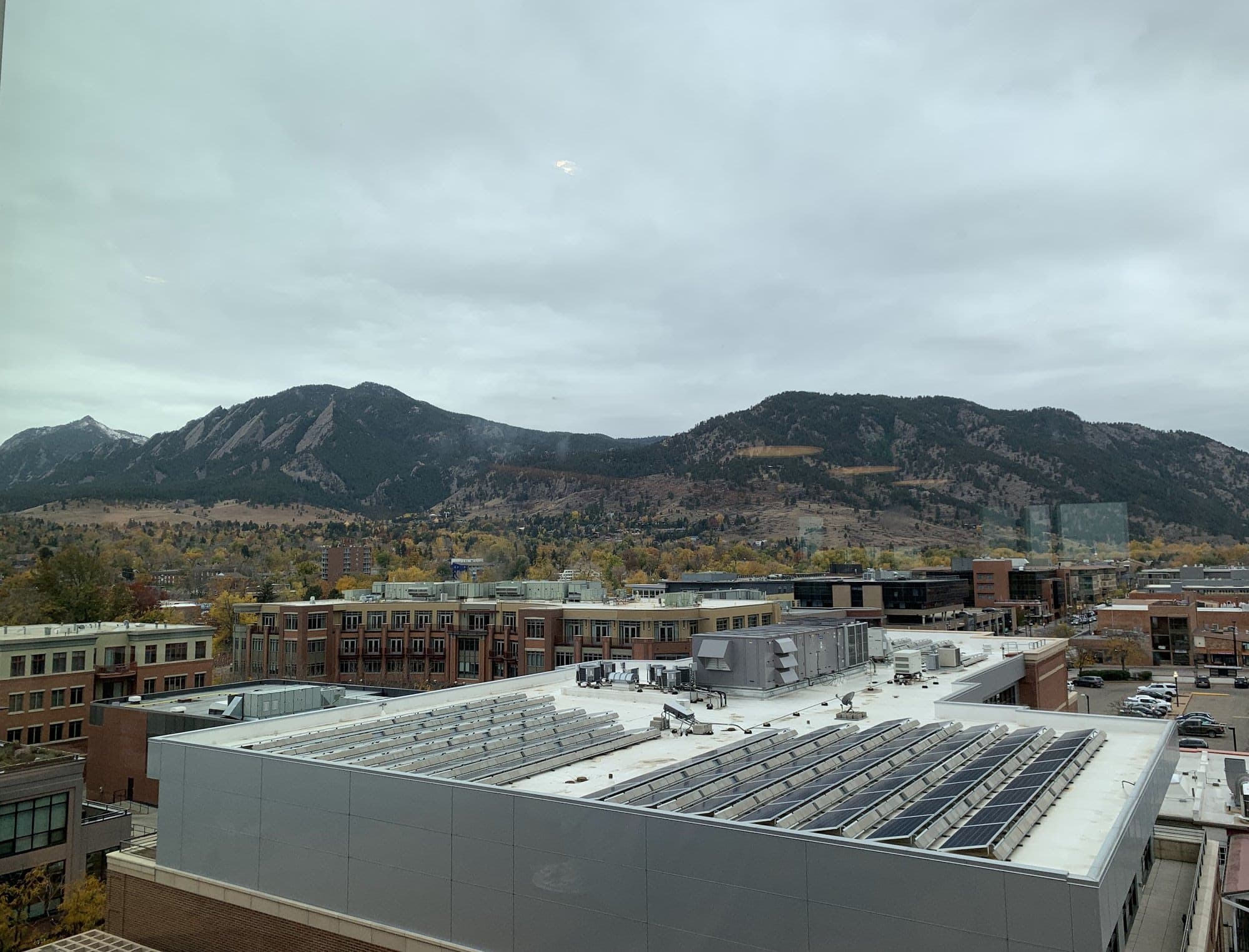 Solar panels on a flat rooftop in Boulder, with brick buildings, autumn trees and the Flatirons under a grey sky