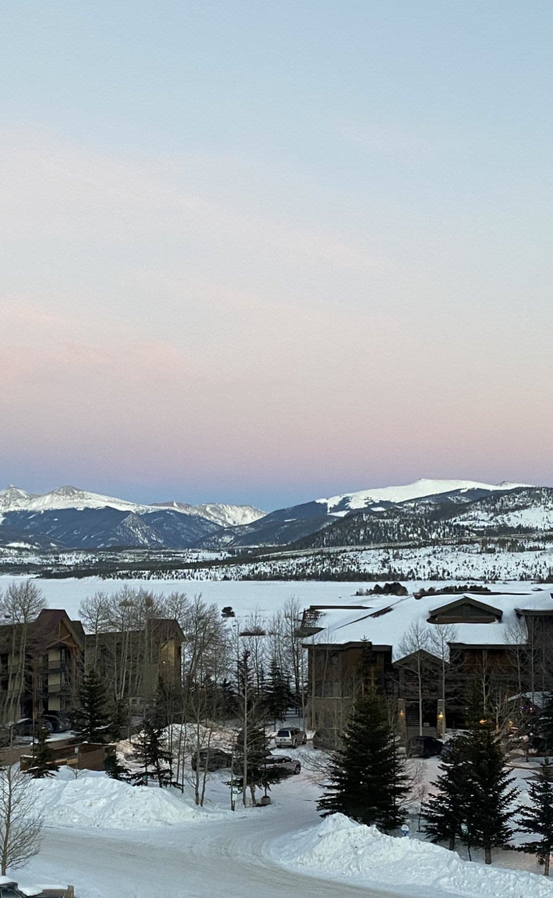 A snowy mountain valley at dusk, pink and blue sky over white peaks, pines and roofs under deep snow