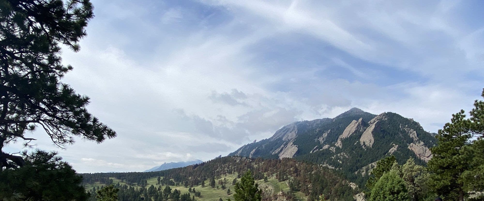 The Flatirons rising above green hills and ponderosa pines under a streaked summer sky
